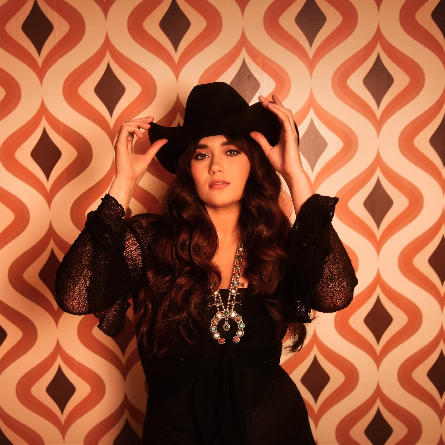 Belles in a black hat against 60s patterned wallpaper